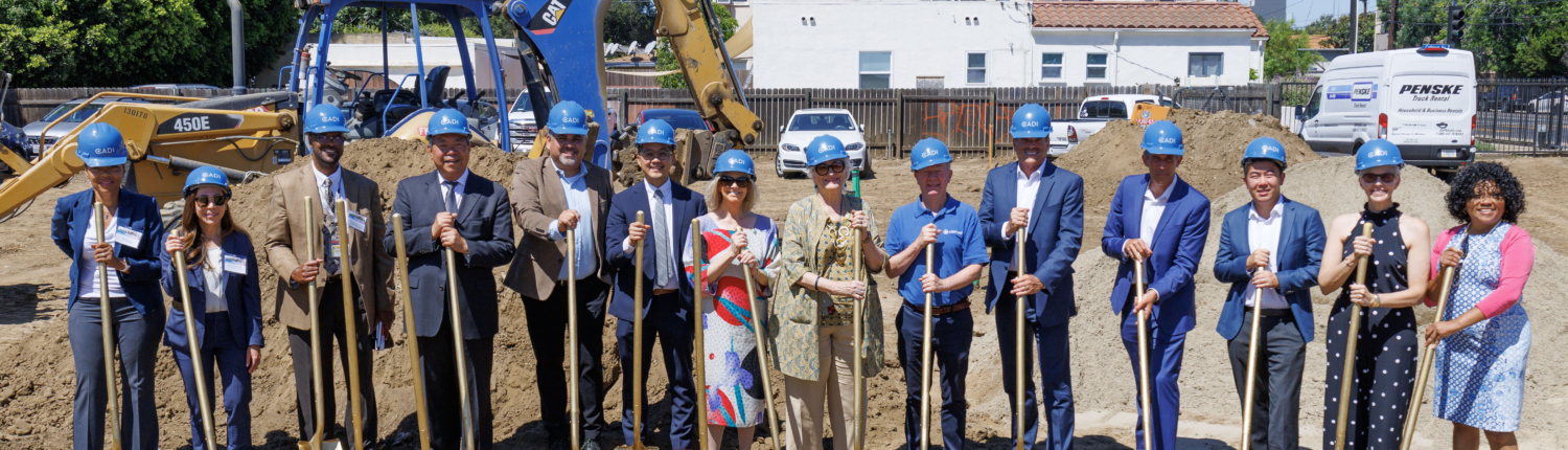 Group of people wearing blue hard hats, holding golden shovels, on dirt lot, with heavy equipment and house in distance behind them.