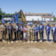 Group of people wearing blue hard hats, holding golden shovels, on dirt lot, with heavy equipment and house in distance behind them.